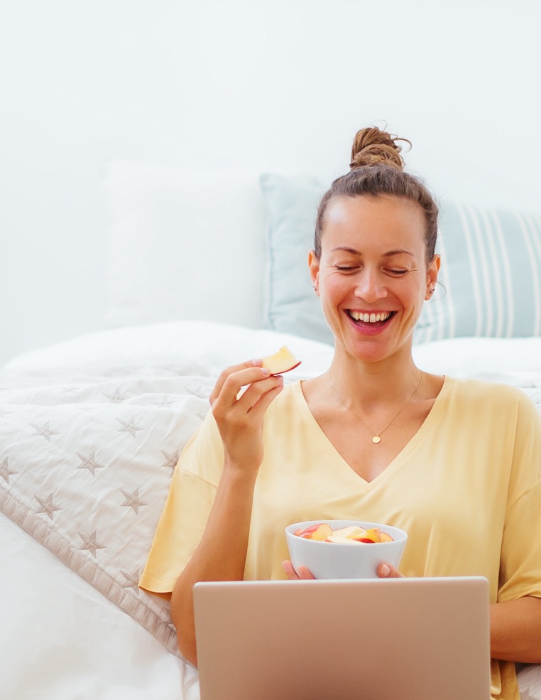 Young woman smiling and looking at her laptop, while snacking on natural chips.